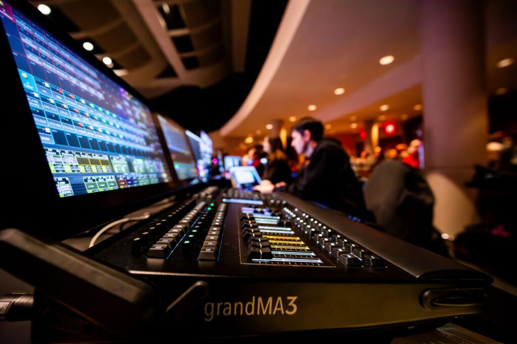 A lighting control console and preview screens at the back of a darkened venue, audience beyond