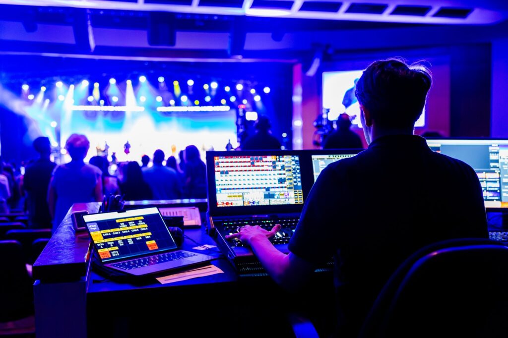 An audiovisual technician works at a lighting and video desk facing a blue-lit stage