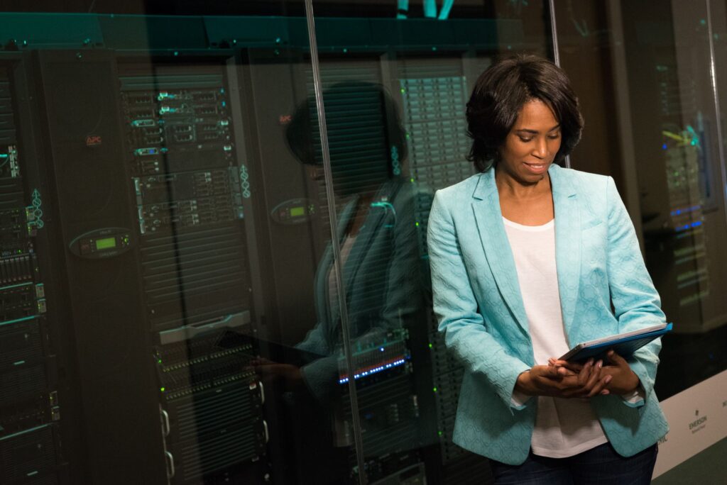 An engineer in a pale green jacket reads a tablet in front of a glass-fronted server cabinet