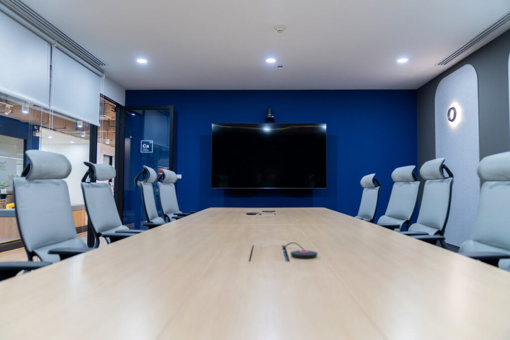 An empty conference room with a long wooden table, grey chairs and a wall-mounted display on a navy feature wall