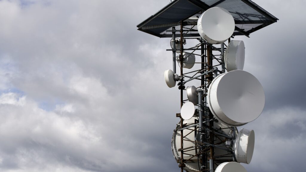 A telecommunications tower hung with white microwave dishes and antennas against a grey cloudy sky