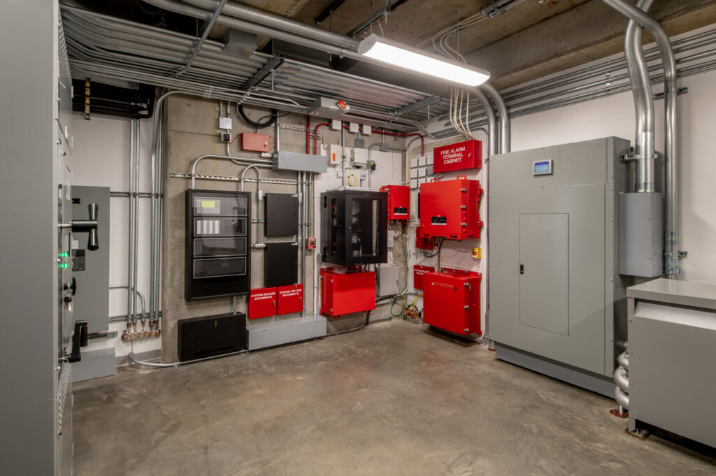 A utility room wall lined with red fire alarm control panels, conduit and electrical enclosures